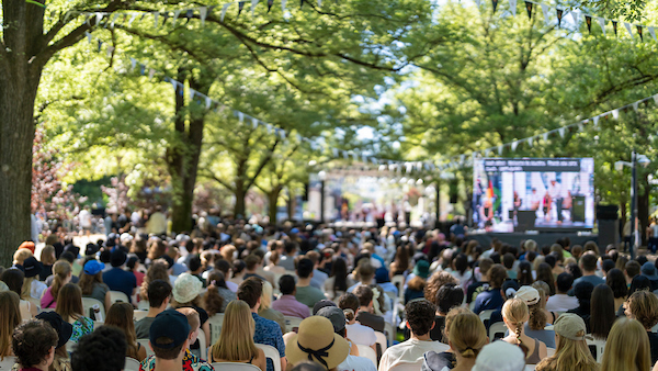 Large crowd seated outdoors beneath trees, watching a talk on a big screen at a public event.